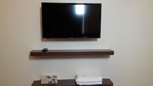 A flat-screen TV mounted on a wall above a wooden shelf. Below the shelf, there is a box labeled "CAREFEEL" and a folded white towel. - De Benz Inn (Malacca City)