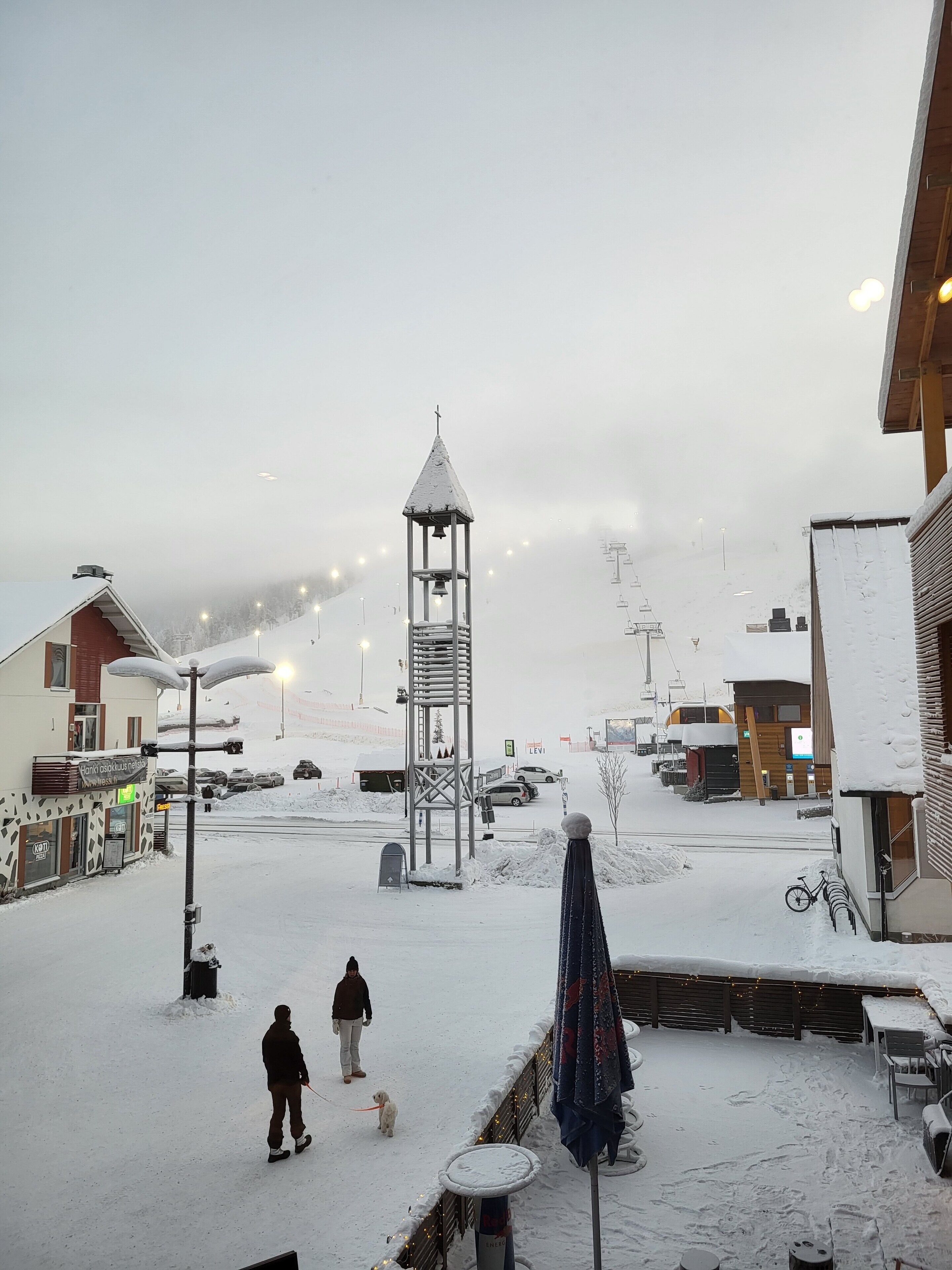 A snowy outdoor area with a bell tower, a covered seating area, and a building with a sign.