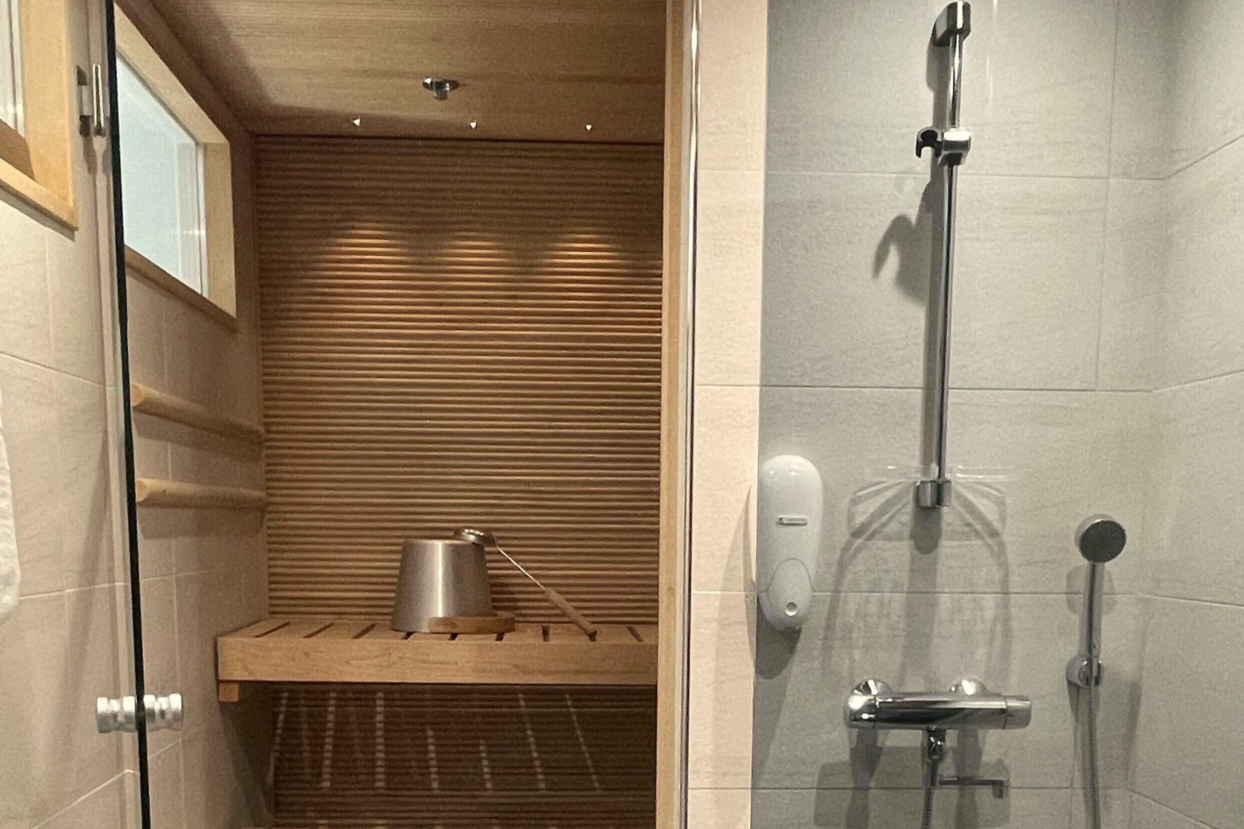 Un espace douche équipé d’un pommeau de douche à main, d’un distributeur de savon mural et d’une étagère en bois avec un récipient en métal.