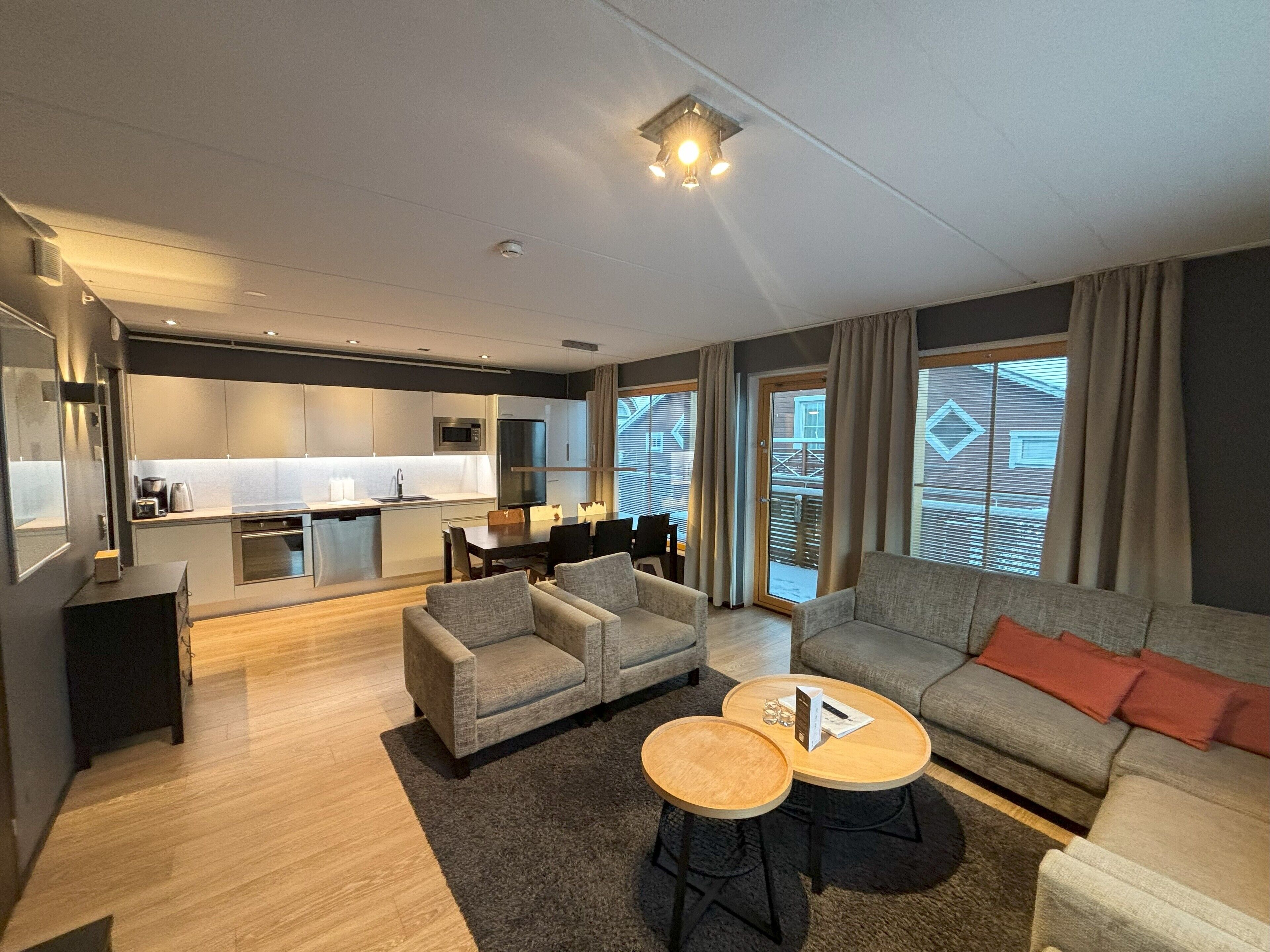 Una sala moderna con sofá, dos sillones, una mesita de centro y una pequeña mesa de comedor.  Hay una zona de cocina con microondas, horno y fregadero. La habitación tiene ventanas grandes con cortinas y vistas al exterior.