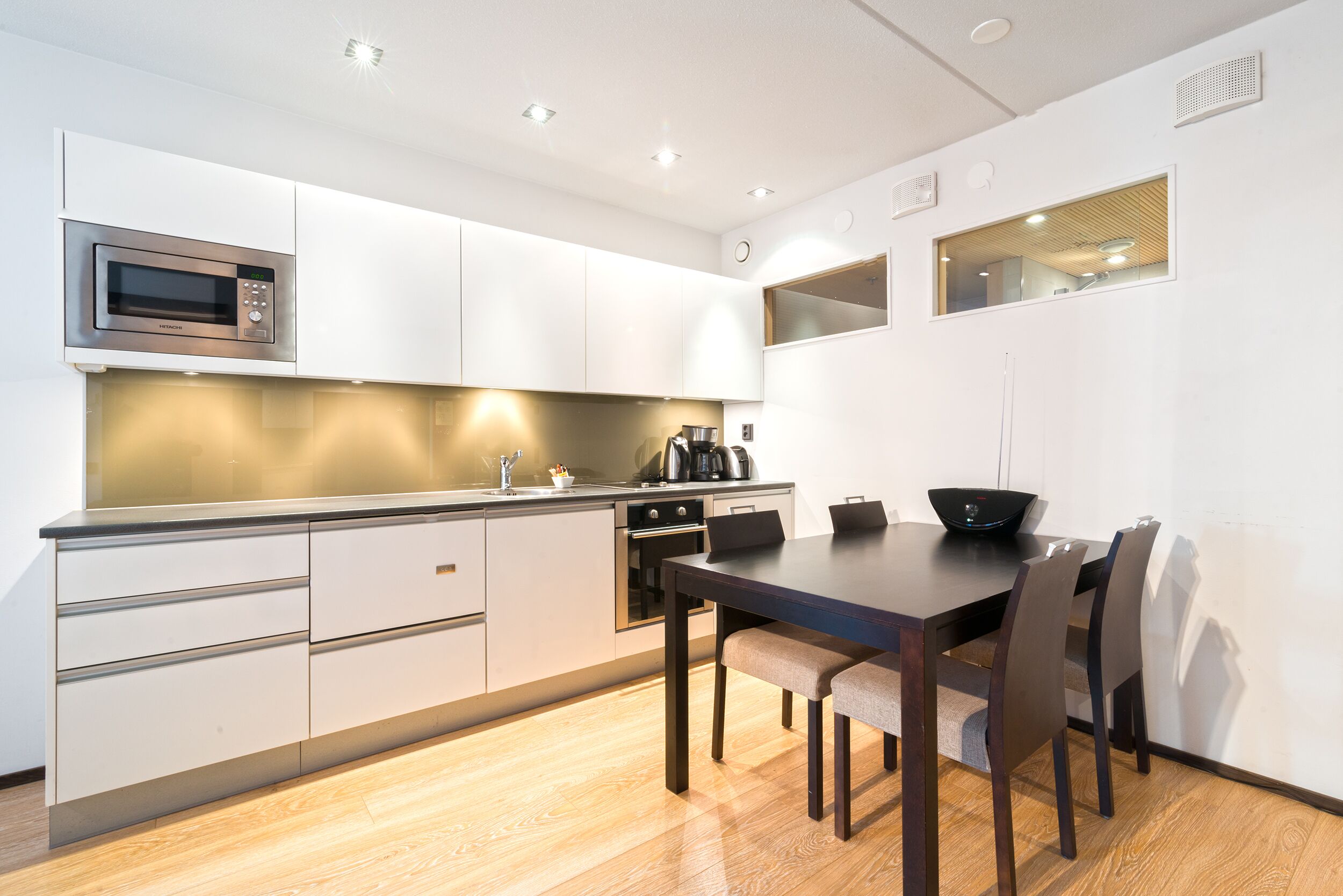 Une cuisine moderne avec des meubles blancs, un four à micro-ondes encastré et un coin repas comprenant une table et des chaises en bois foncé.