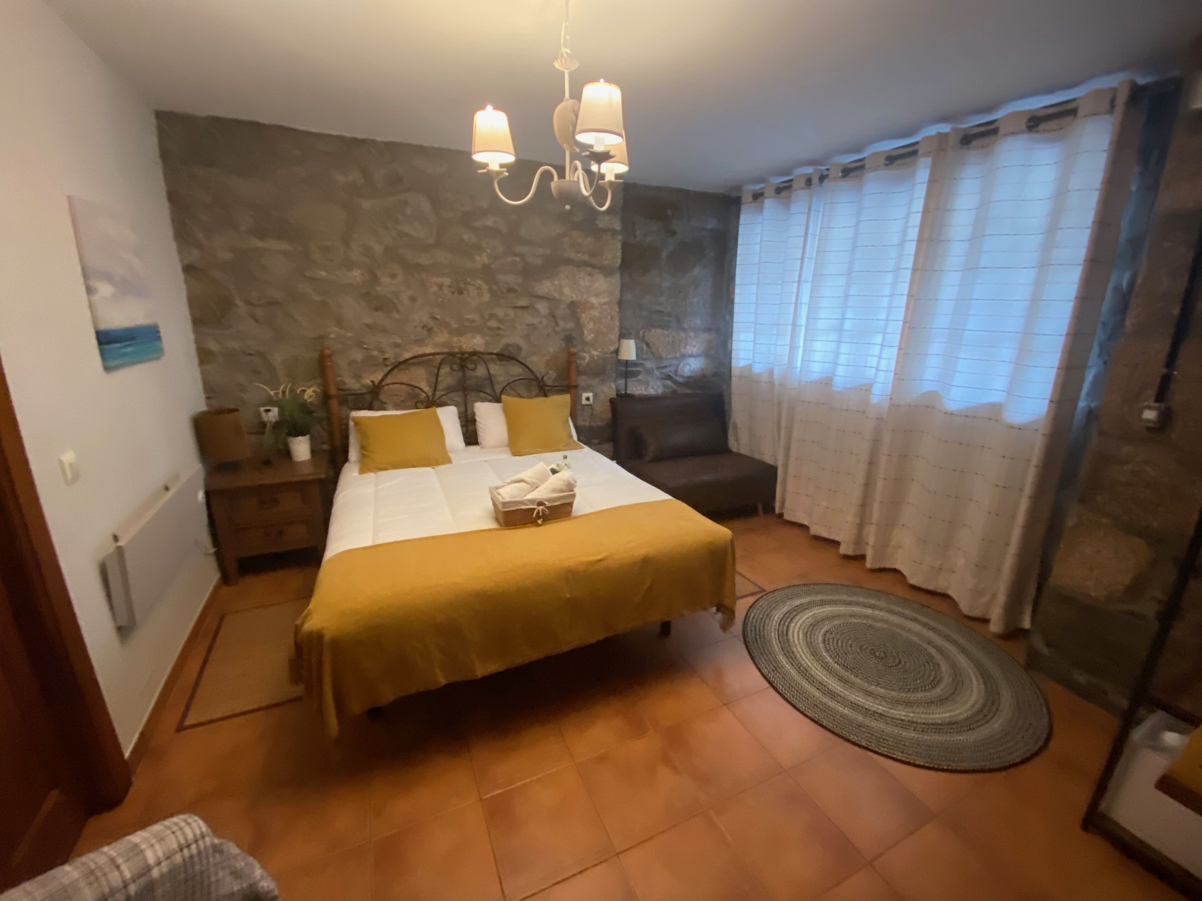 A bedroom with a bed, a nightstand, a dresser, a chair, a window with curtains, and a chandelier.
