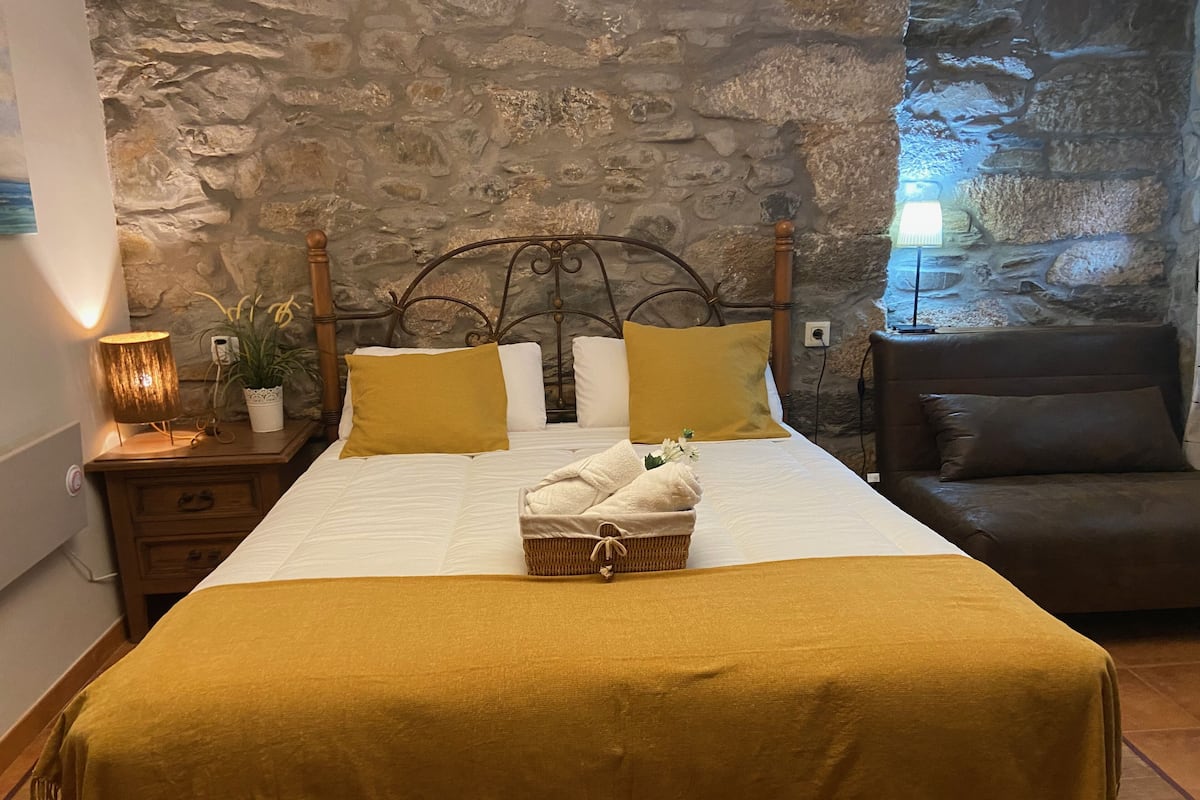 Una cama bien hecha con edredón blanco y dos almohadas color mostaza, una cesta de mimbre con toallas, una mesita de noche con lámpara, y una pared de piedra de fondo.