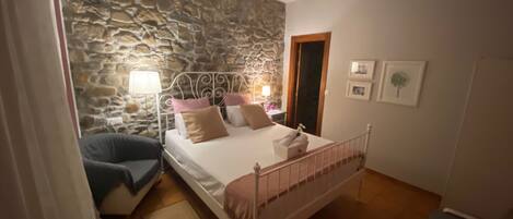 Un dormitorio con pared de piedra, cama con almohadas, mesita de noche con lámpara y una silla.