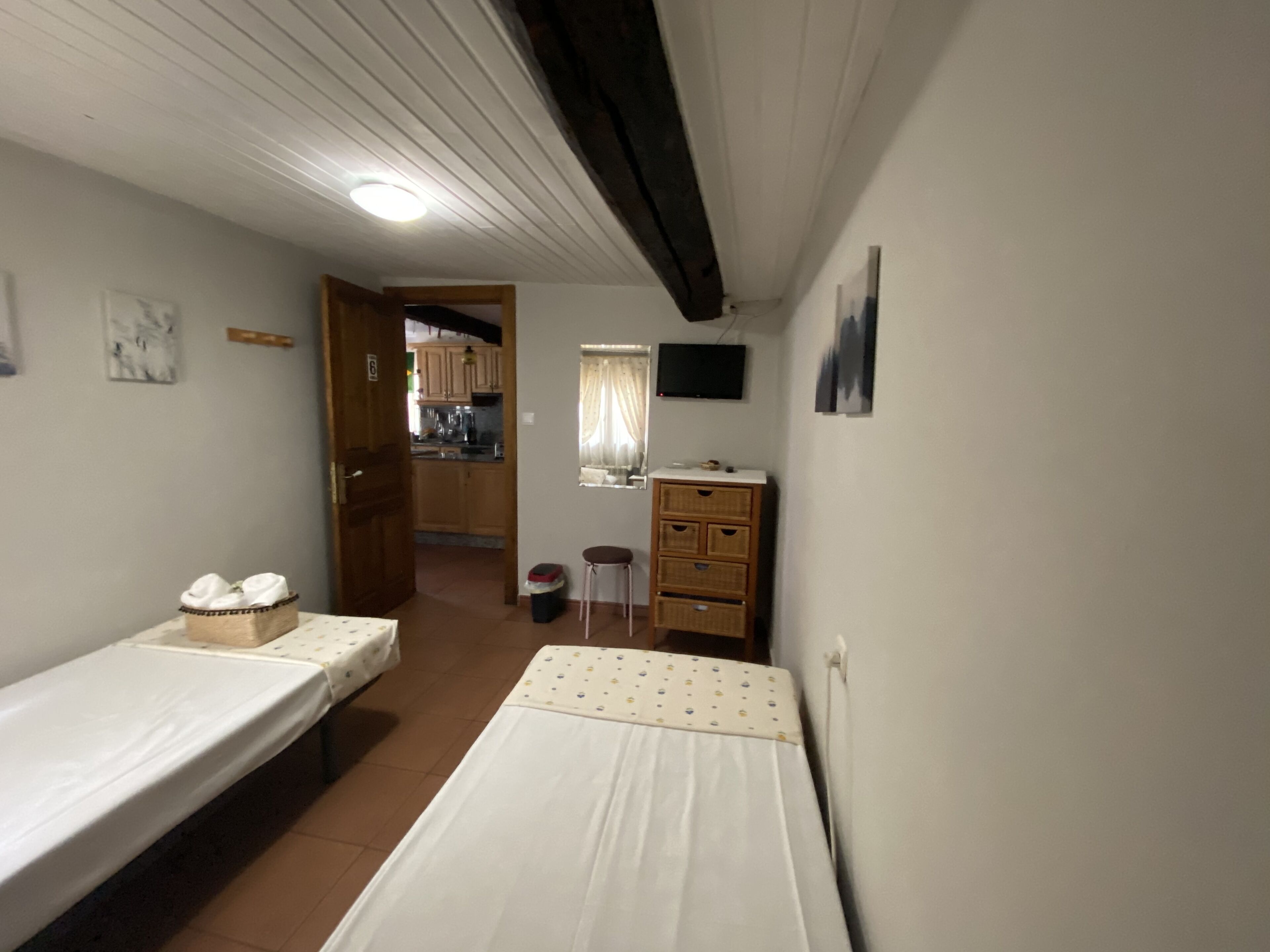 Een kleine, eenvoudige kamer met twee eenpersoonsbedden, een houten dressoir, een televisie en een keukenruimte zichtbaar door een geopende deur.