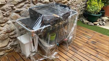 Una parrilla para barbacoa envuelta en plástico, frente a una pared de piedra y una terraza de madera.