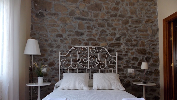 Una cama bien hecha con sábanas y almohadas blancas, y un muro de piedra al fondo, además de dos mesitas de noche con lámparas.
