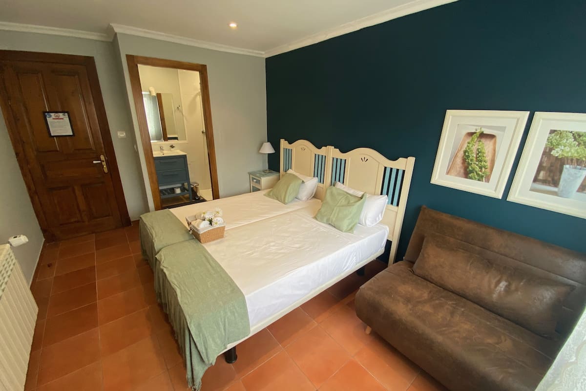 Un dormitorio con cama, un sillón marrón, una puerta de madera, un espejo de baño y un cuadro en la pared.