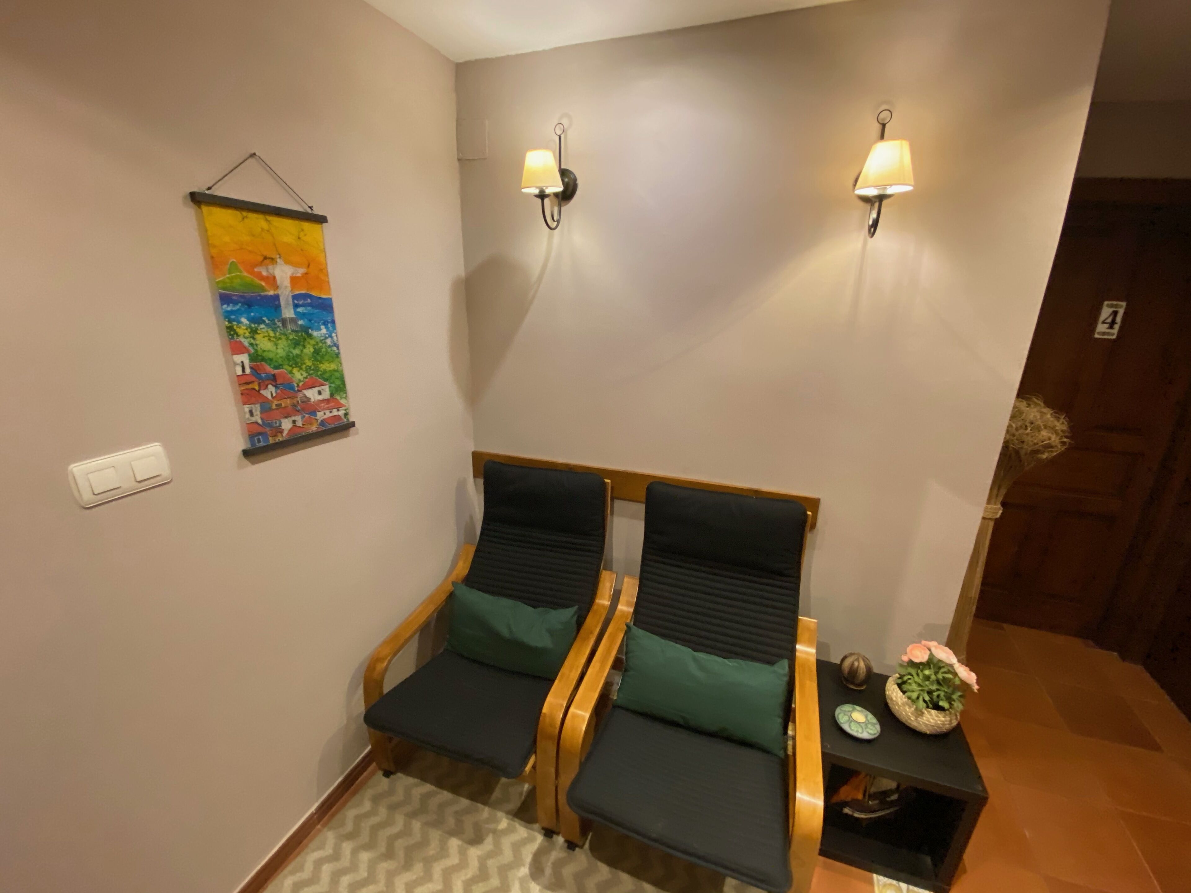 Una habitación con dos sillas de madera con cojines negros, una lámpara de pared y un cuadro de un paisaje.