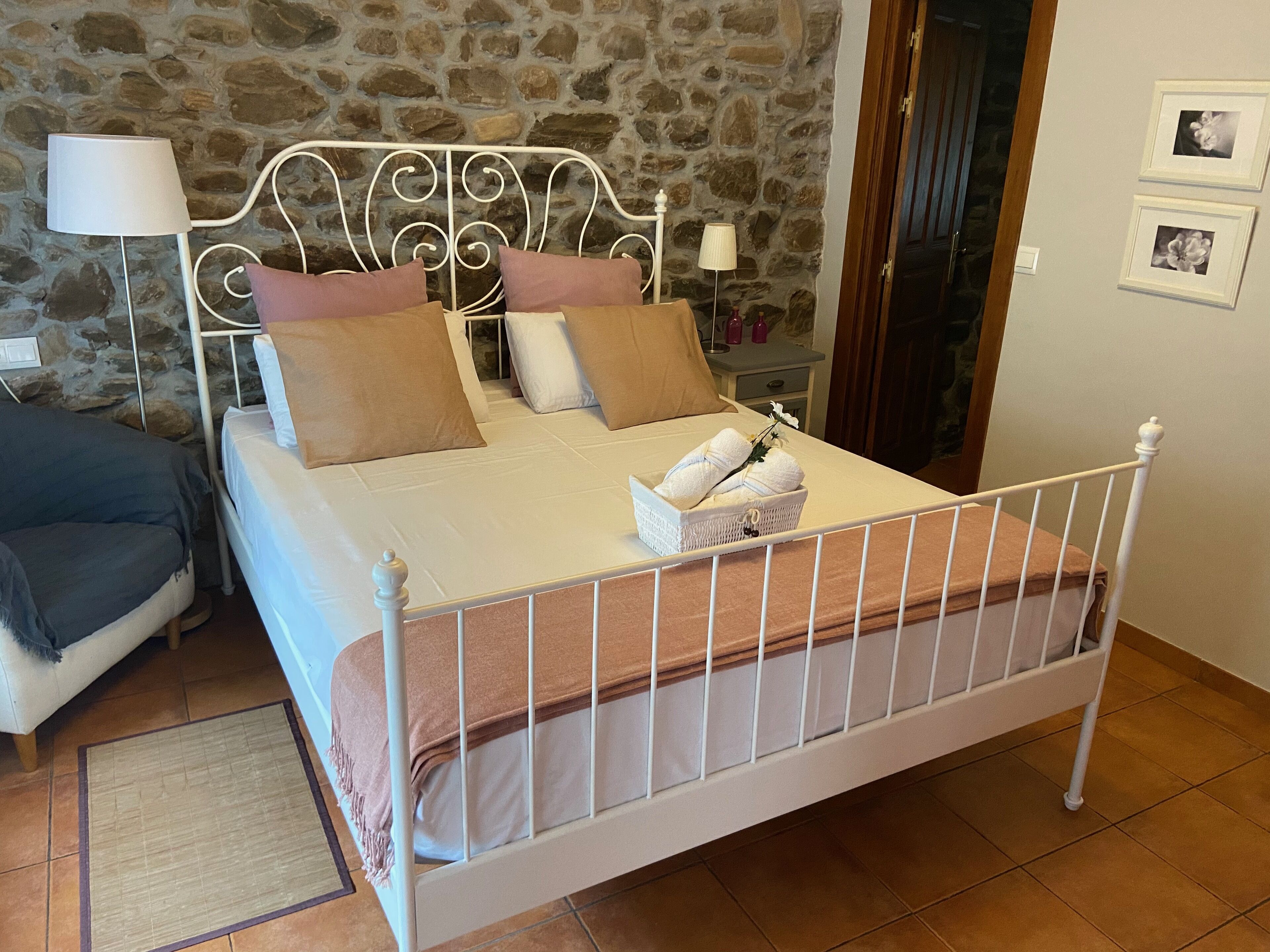 Una cama con estructura metálica, ropa de cama blanca y una manta marrón. Hay una mesita de noche con una lámpara y una pequeña mesa con un jarrón. La habitación tiene paredes de piedra y una puerta de madera.