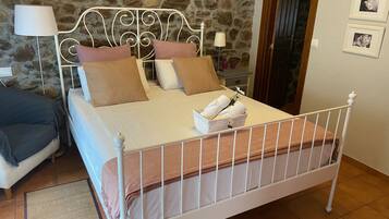Una cama con estructura metálica, ropa de cama blanca y una manta marrón. Hay una mesita de noche con una lámpara y una pequeña mesa con un jarrón. La habitación tiene paredes de piedra y una puerta de madera.