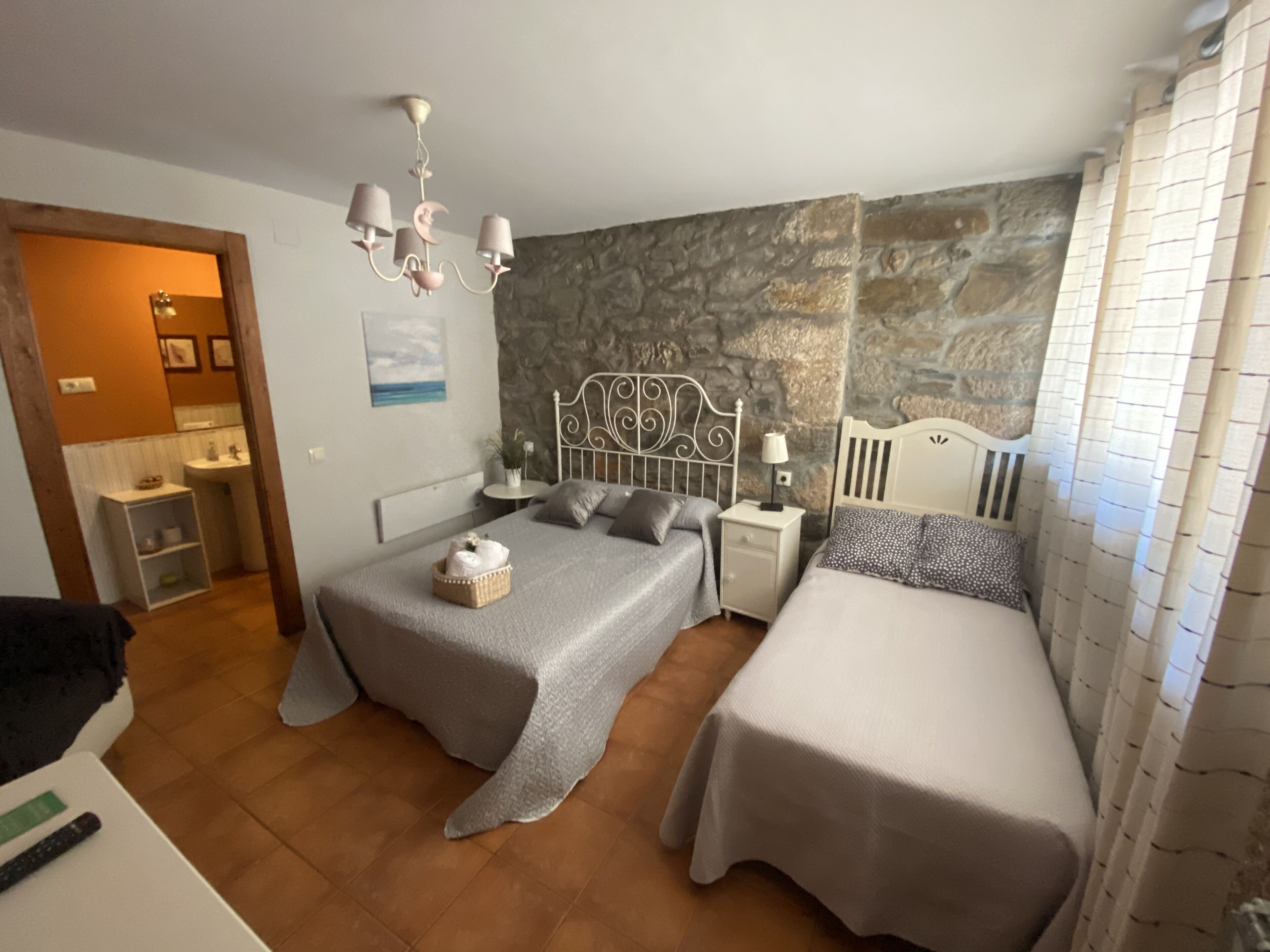 Un dormitorio con dos camas, una pared de piedra, una araña y un baño visible a través de una puerta abierta.