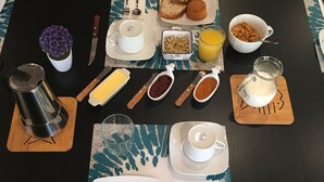Una mesa de desayuno servida con platos, cubiertos, una cafetera, una jarra de leche, un tazón de cereal y un vaso de jugo de naranja, además de una pequeña planta en maceta.