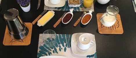 A breakfast table set with plates, cutlery, a coffee pot, a jug of milk, a bowl of cereal, a glass of orange juice, and a small potted plant.