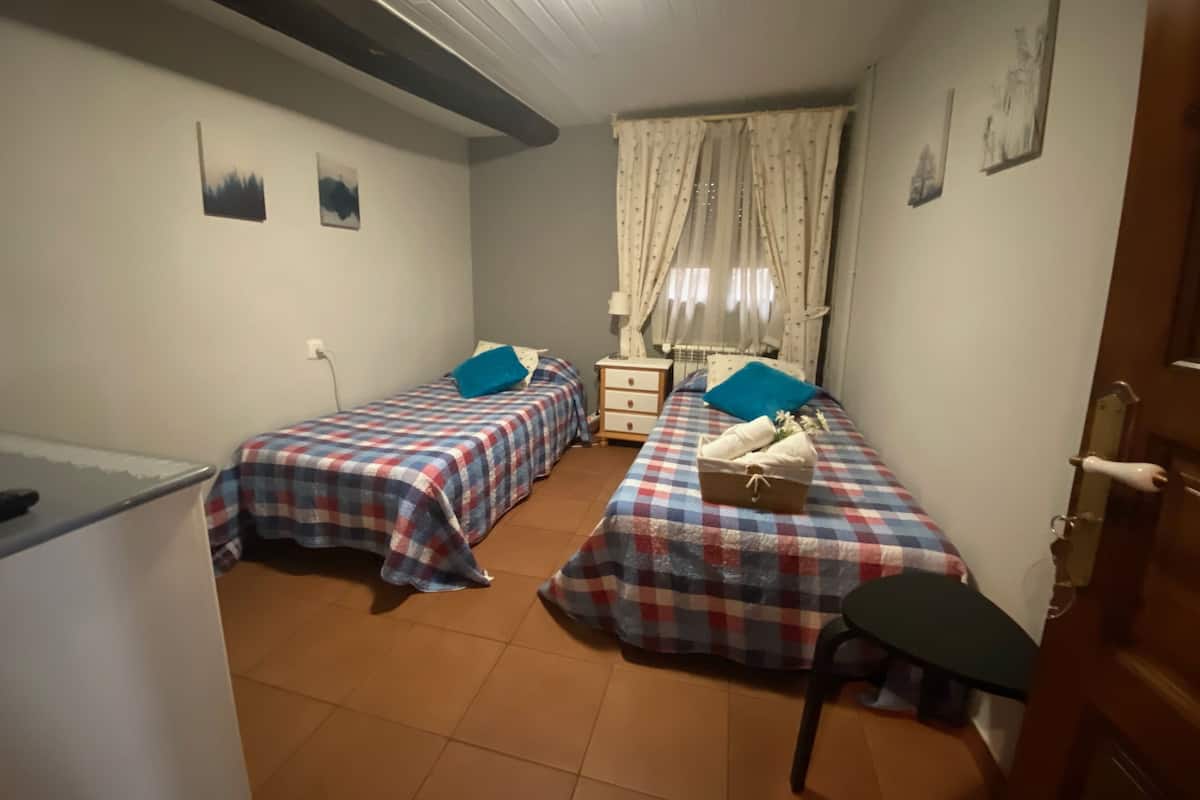 Una habitación con dos camas, una mesita, una silla, una lámpara y un ventanal con cortinas.