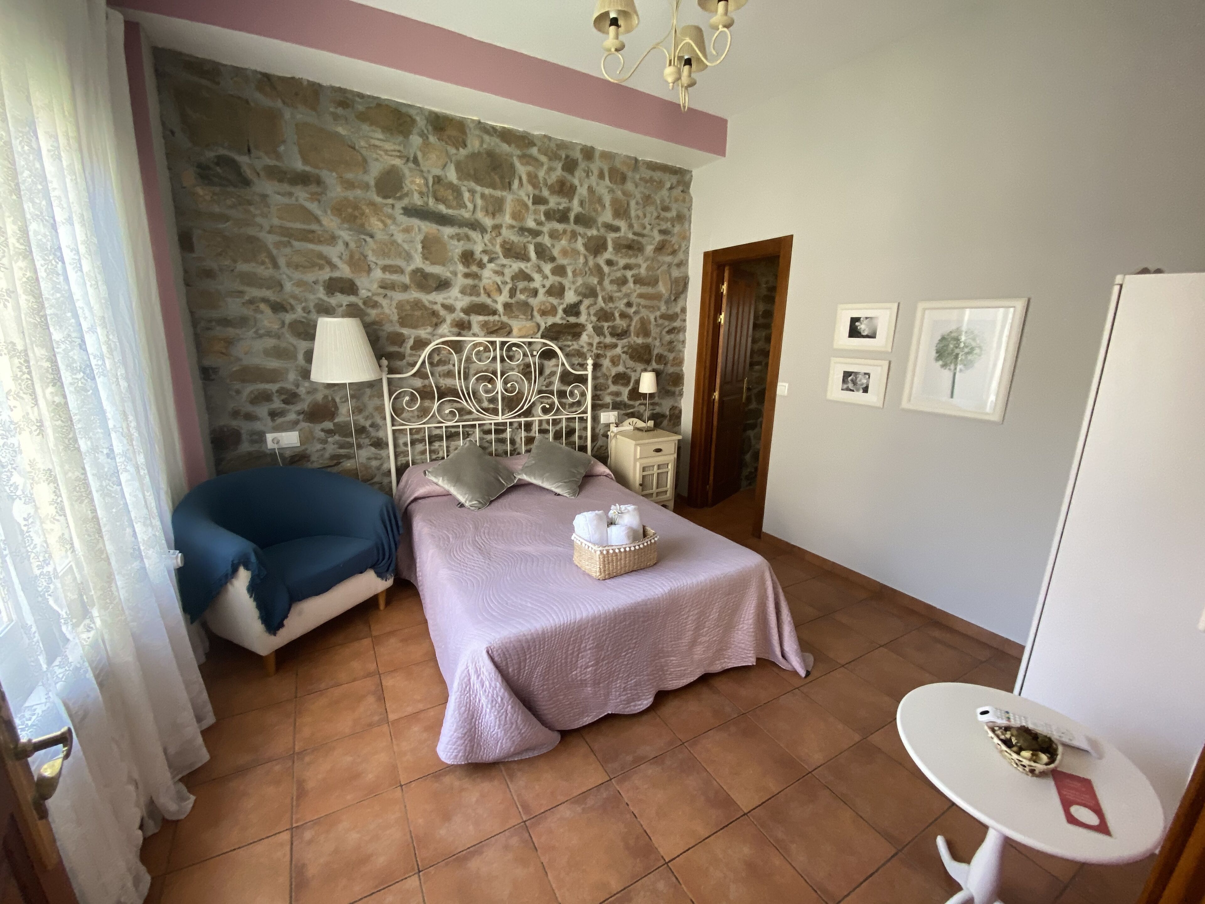 Un dormitorio con pared de piedra, una cama con cabecera, una mesita de noche con lámpara, una pequeña mesa con una cesta y un armario blanco.