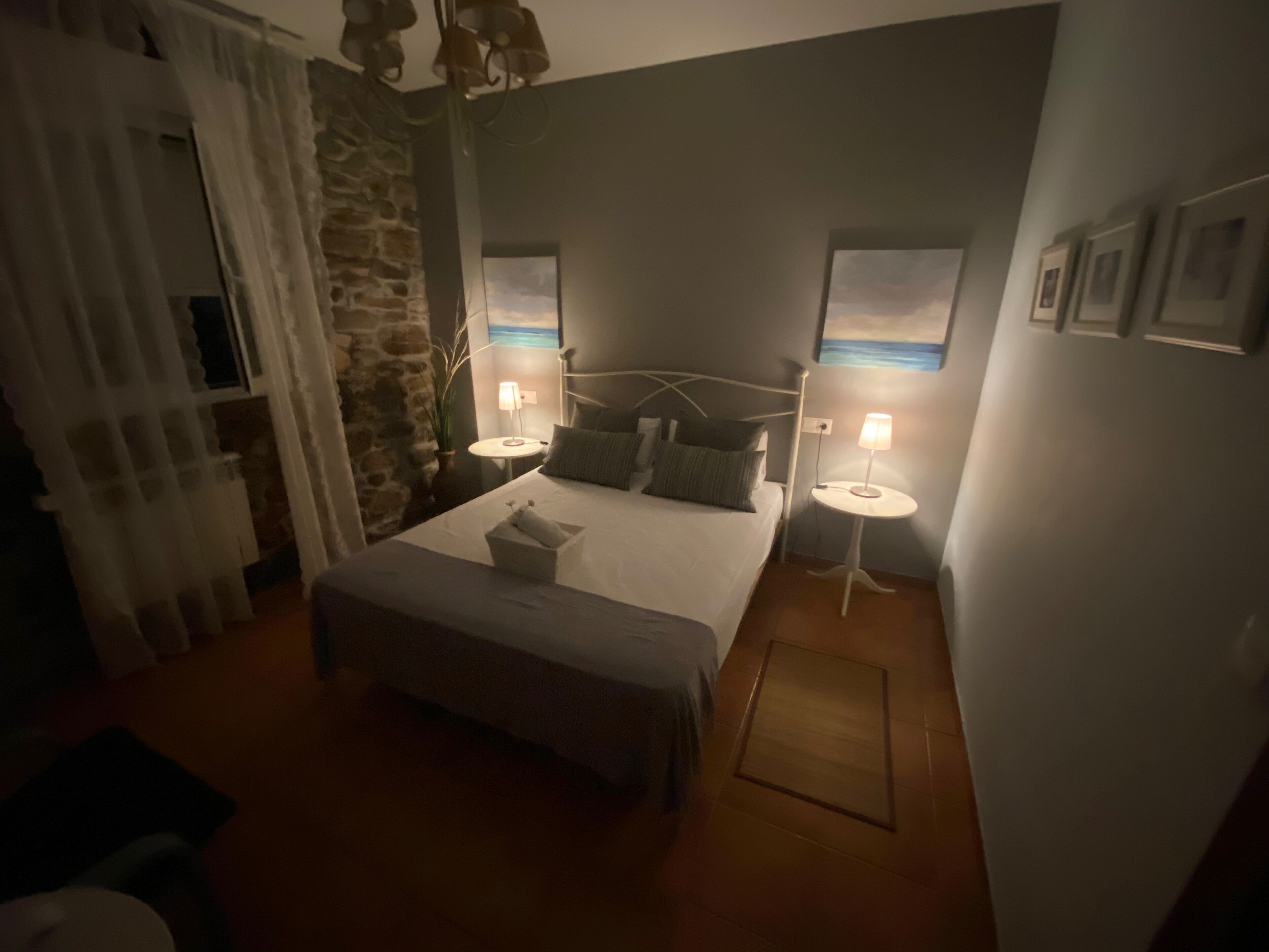 Un dormitorio con cama, dos mesitas de noche con lámparas, pared de piedra y vista al mar.