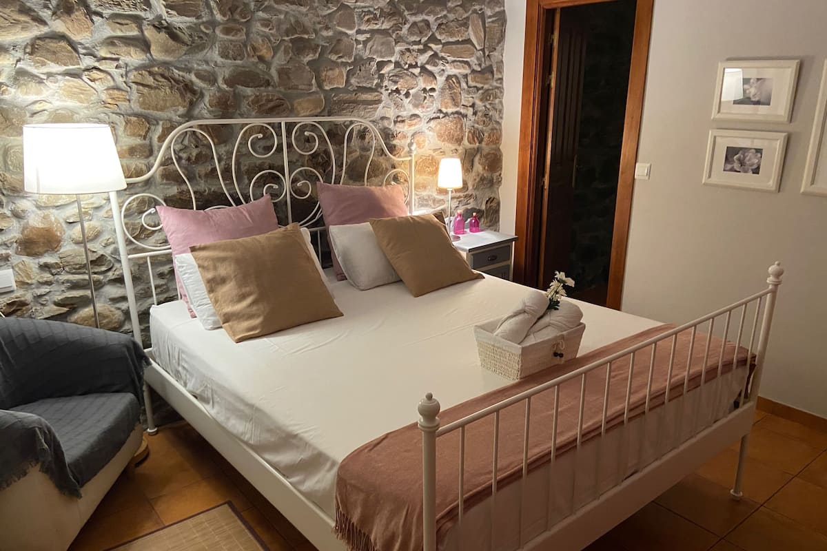 Un dormitorio con pared de piedra, una cama con almohadas, una mesita de noche con lámpara y una silla.