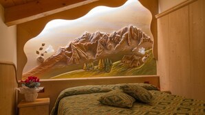 A bedroom with a mountain landscape mural on the wall, a wooden bed frame, and a nightstand with a vase of flowers. - Hotel Orchidea (Vermiglio)