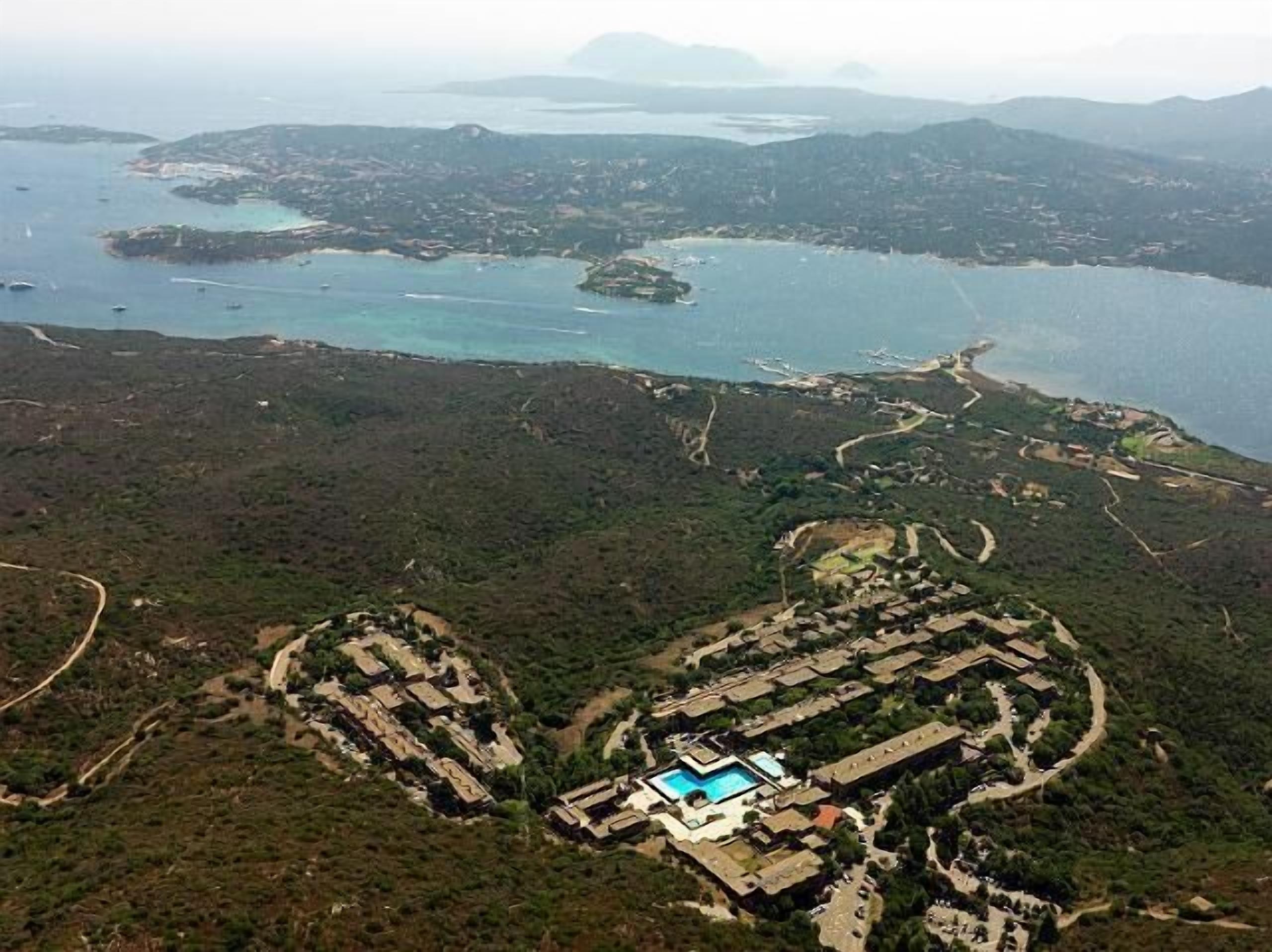 Vista aérea de un complejo turístico frente al mar con varias construcciones, una piscina y un paisaje natural circundante.