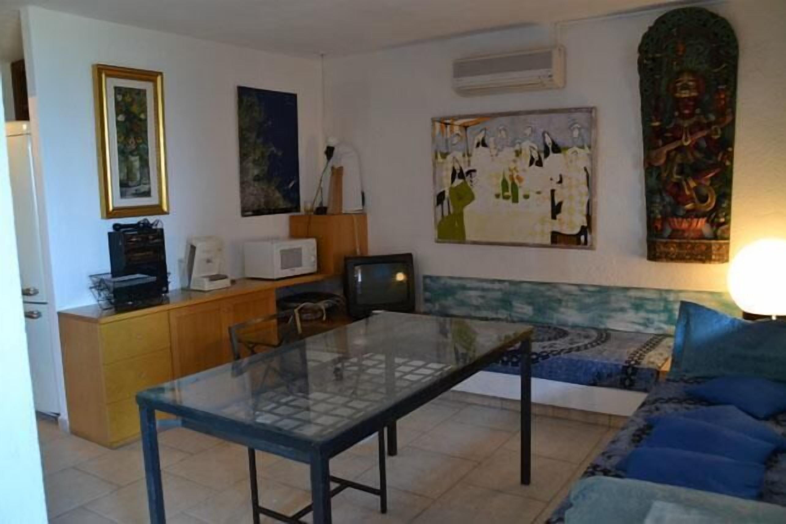 Ein Wohnzimmer mit Glastisch, einer Couch und einem Fernseher. An den Wänden befinden sich Gemälde und eine kleine Vitrine mit einer Mikrowelle.
