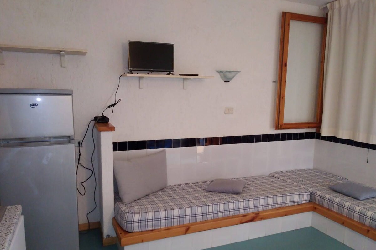 A compact room with a bed, a television on a shelf, a refrigerator, and a window with curtains.
