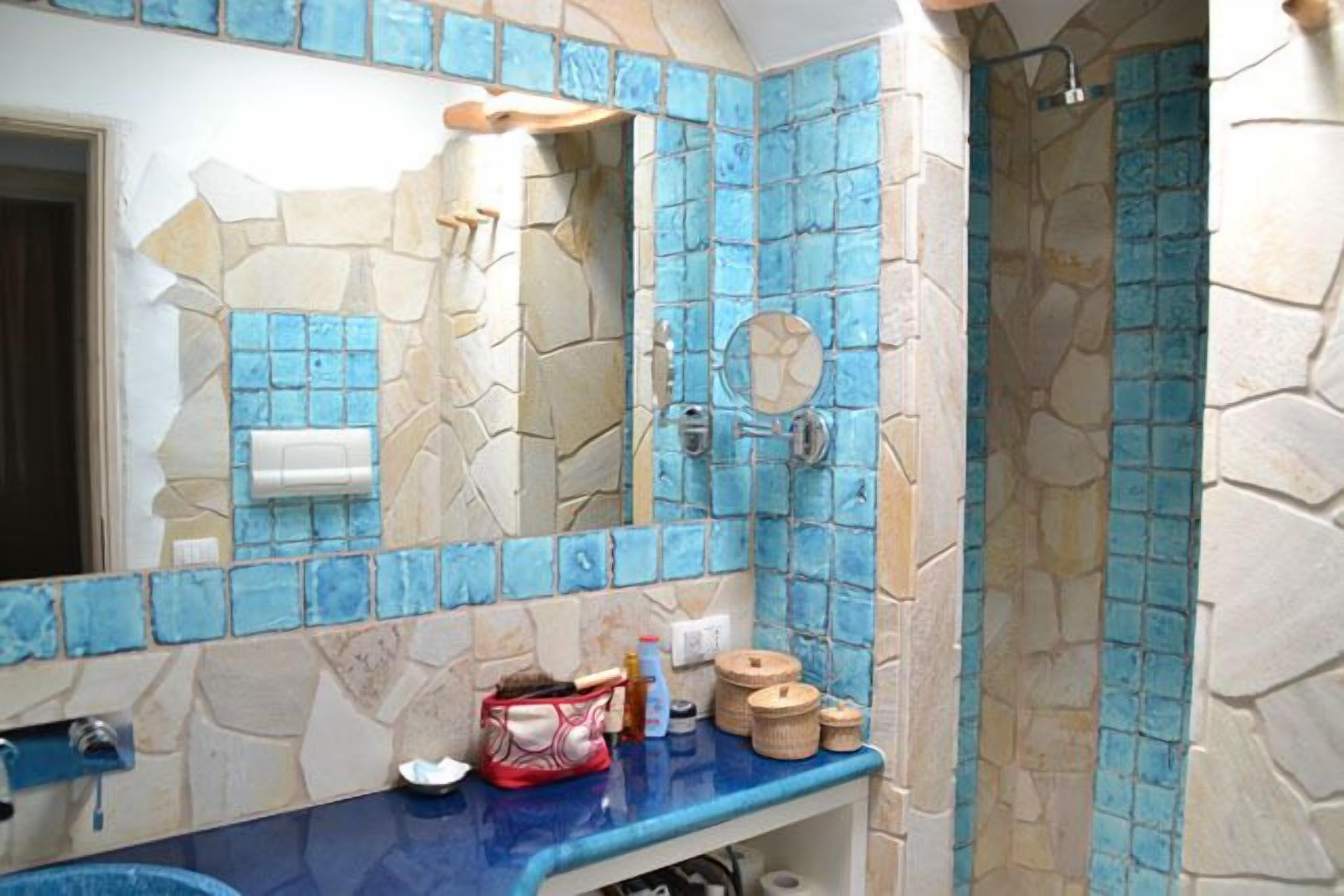 A bathroom with a stone and blue mosaic tile design, a sink with toiletries, and a shower area.