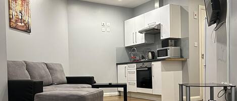 A modern living room with a grey sofa, a coffee table, and a kitchen area with white cabinets and appliances.
