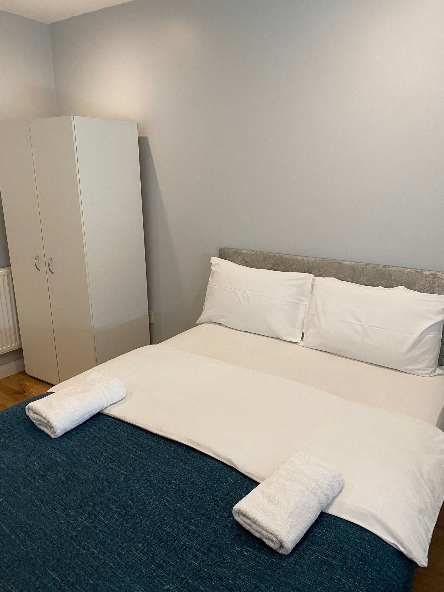 Un lit bien fait, avec du linge de lit blanc et deux serviettes, une tête de lit grise et une armoire blanche, dans une pièce aux murs de couleur claire.