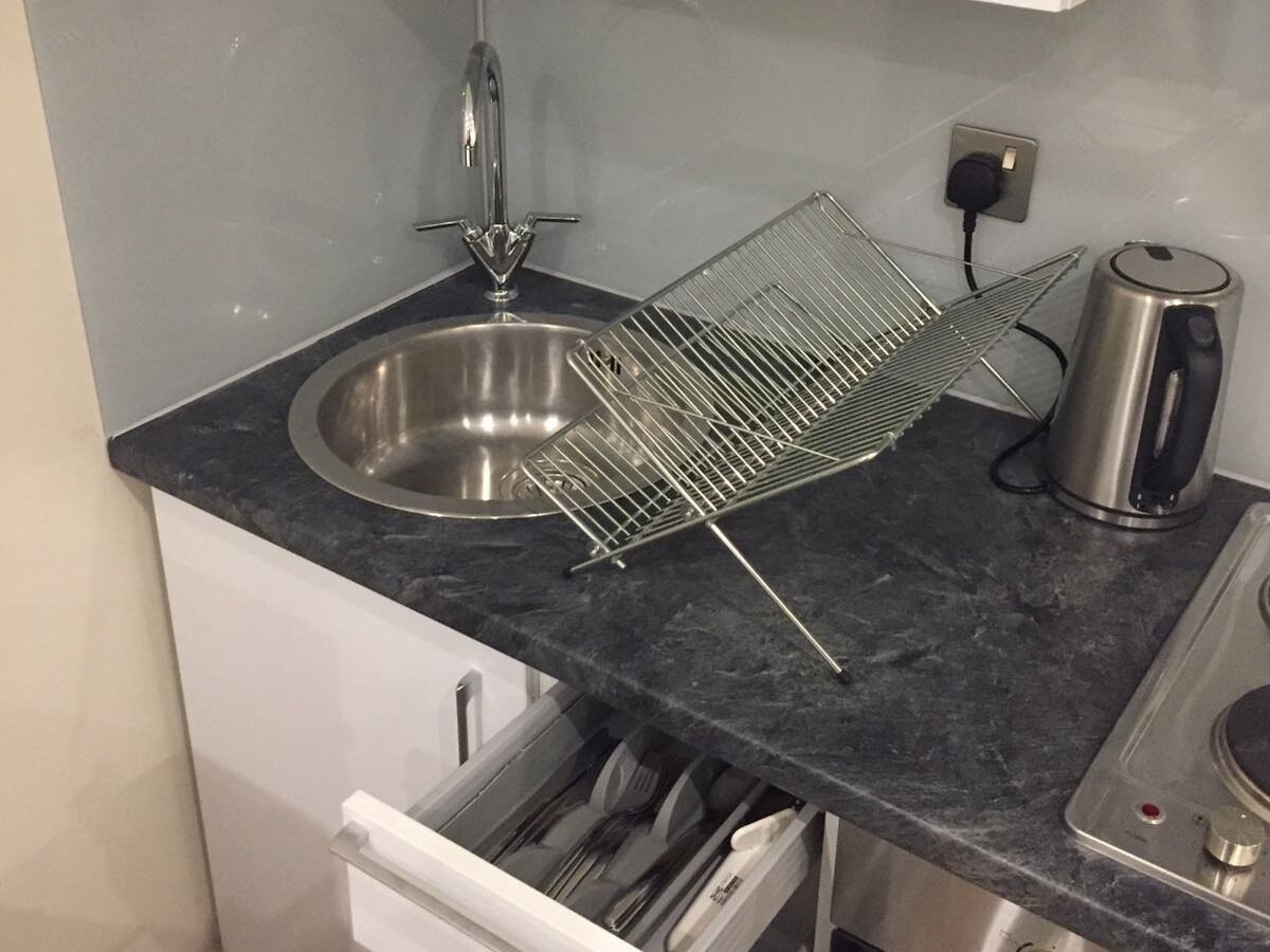 A kitchen with a stainless steel sink, a kettle, and a dish rack on a dark countertop.