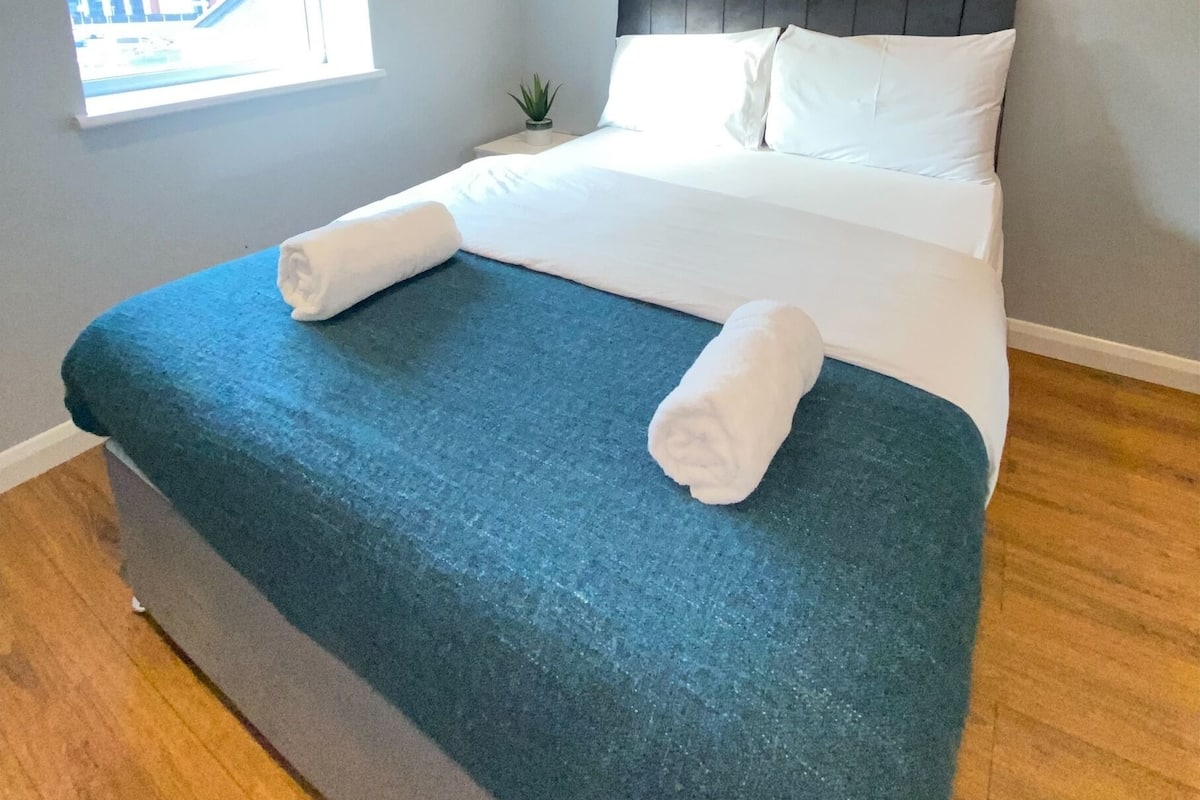A neatly made bed with white linens and a teal blanket, a small potted plant on the nightstand, and a window with a view of buildings outside.