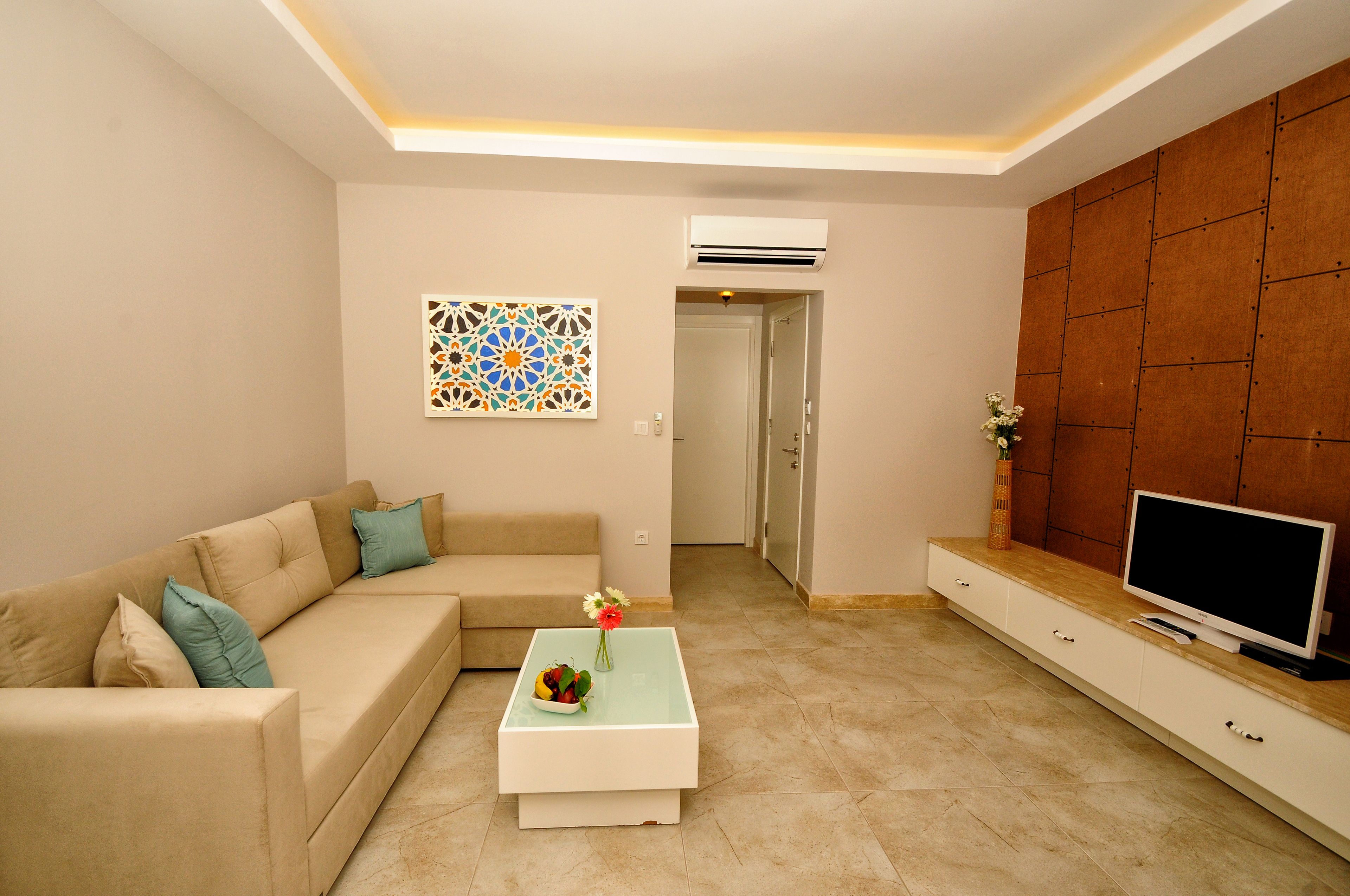 A living room with a beige sofa, a coffee table with a bowl of fruit and flowers, a flat-screen TV on a wooden cabinet, and a decorative wall piece.