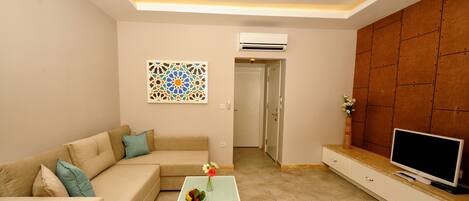 A living room with a beige sofa, a coffee table with a bowl of fruit and flowers, a flat-screen TV on a wooden cabinet, and a decorative wall piece.
