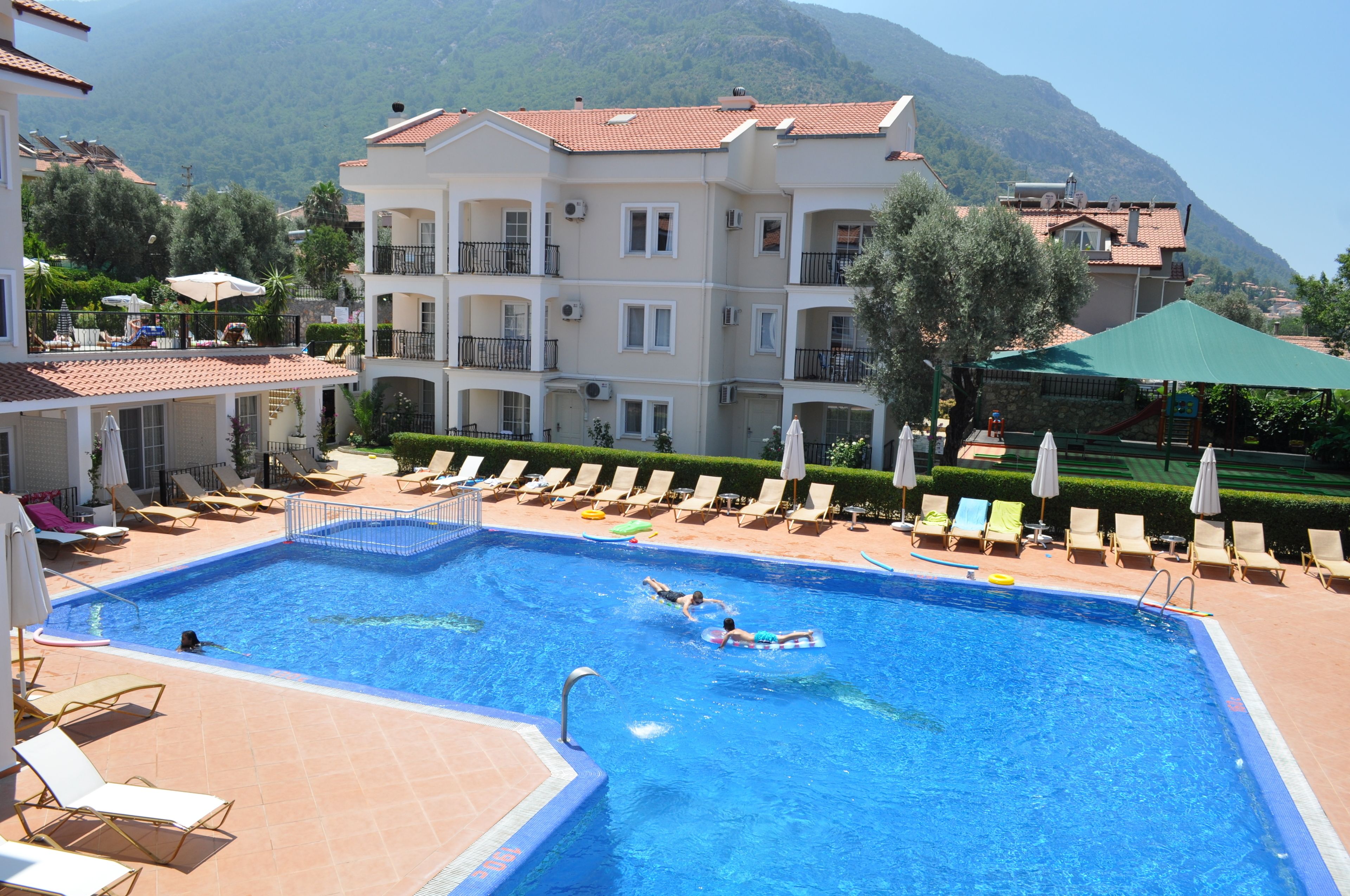 A swimming pool with lounge chairs and umbrellas, surrounded by buildings with balconies and a mountain in the background.