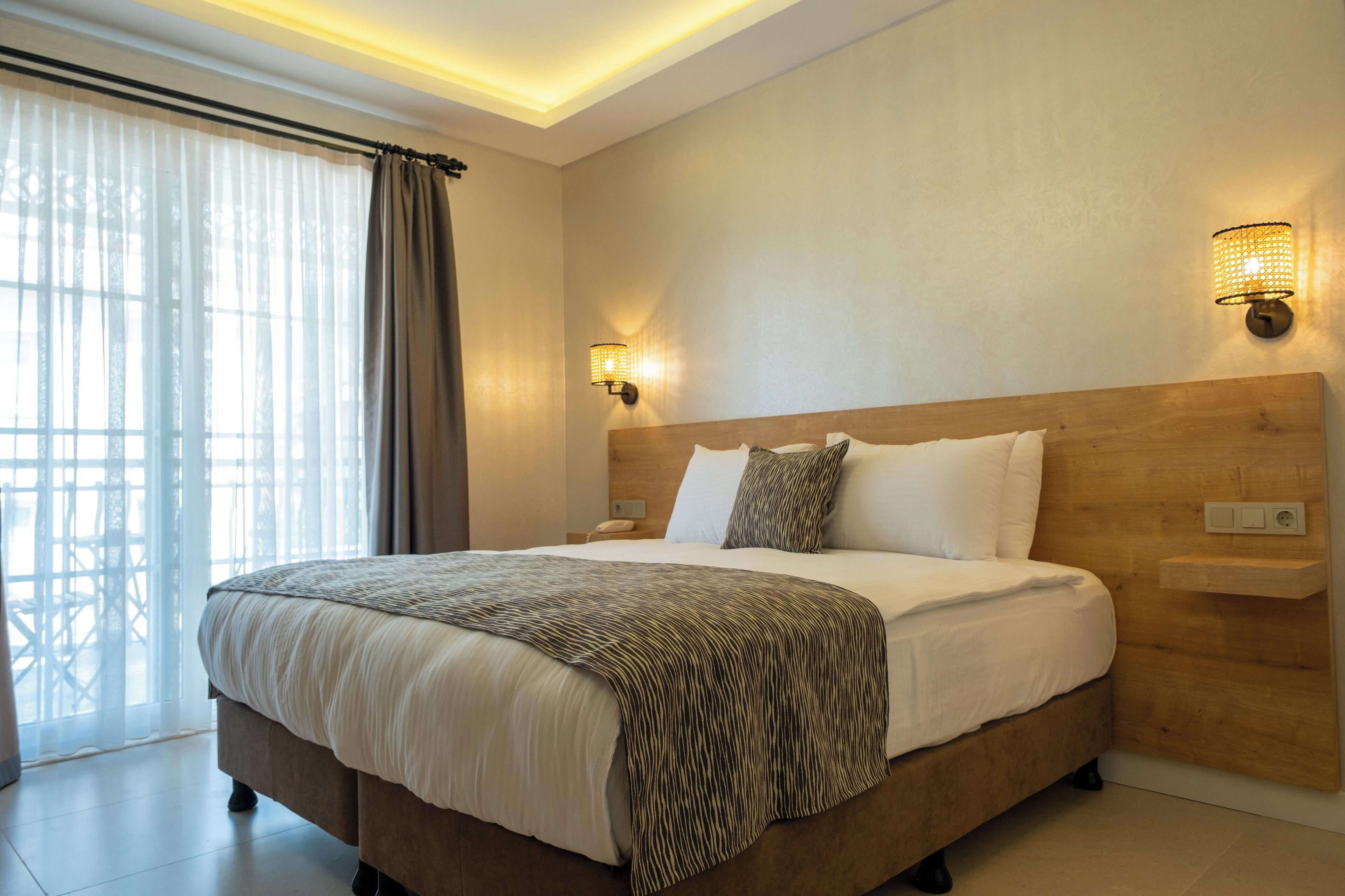 A modern hotel room with a large bed, wooden headboard, two bedside lamps, and a view of the outdoors through sheer curtains.