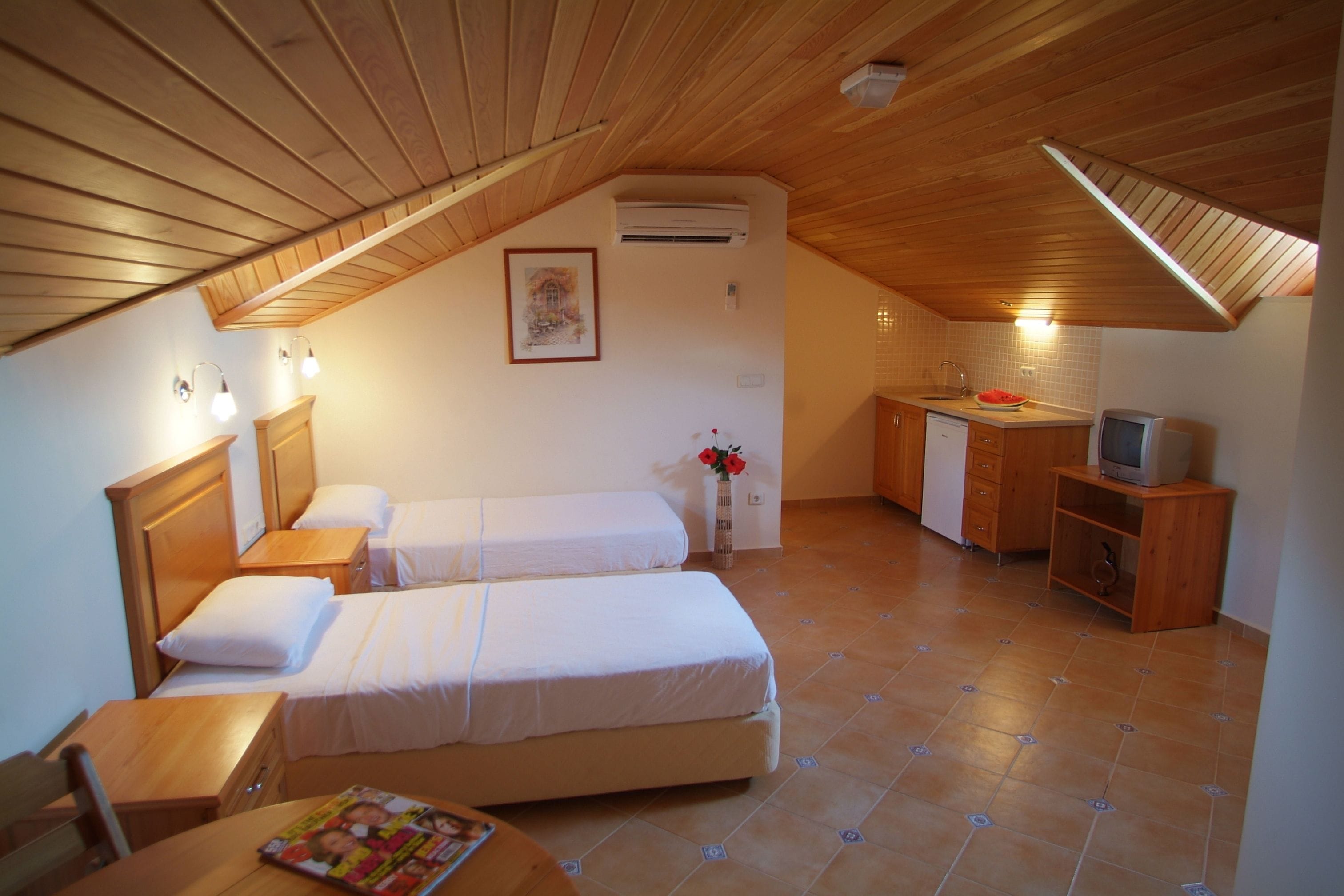 A cozy attic bedroom with two beds, a small desk, a TV, and a kitchenette.