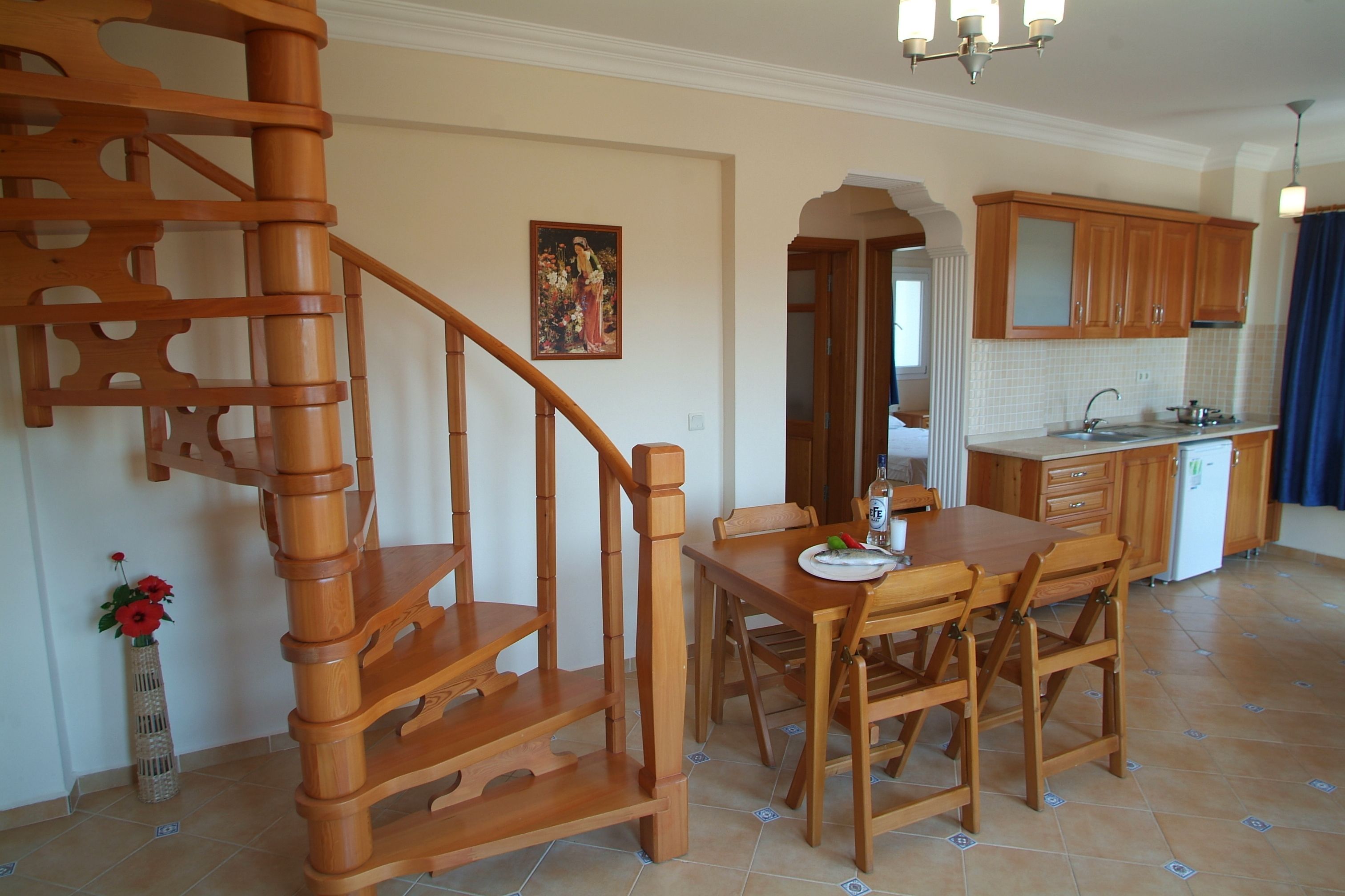 A wooden staircase, a dining table with chairs, a kitchen area with wooden cabinets, and a vase with flowers on the floor.