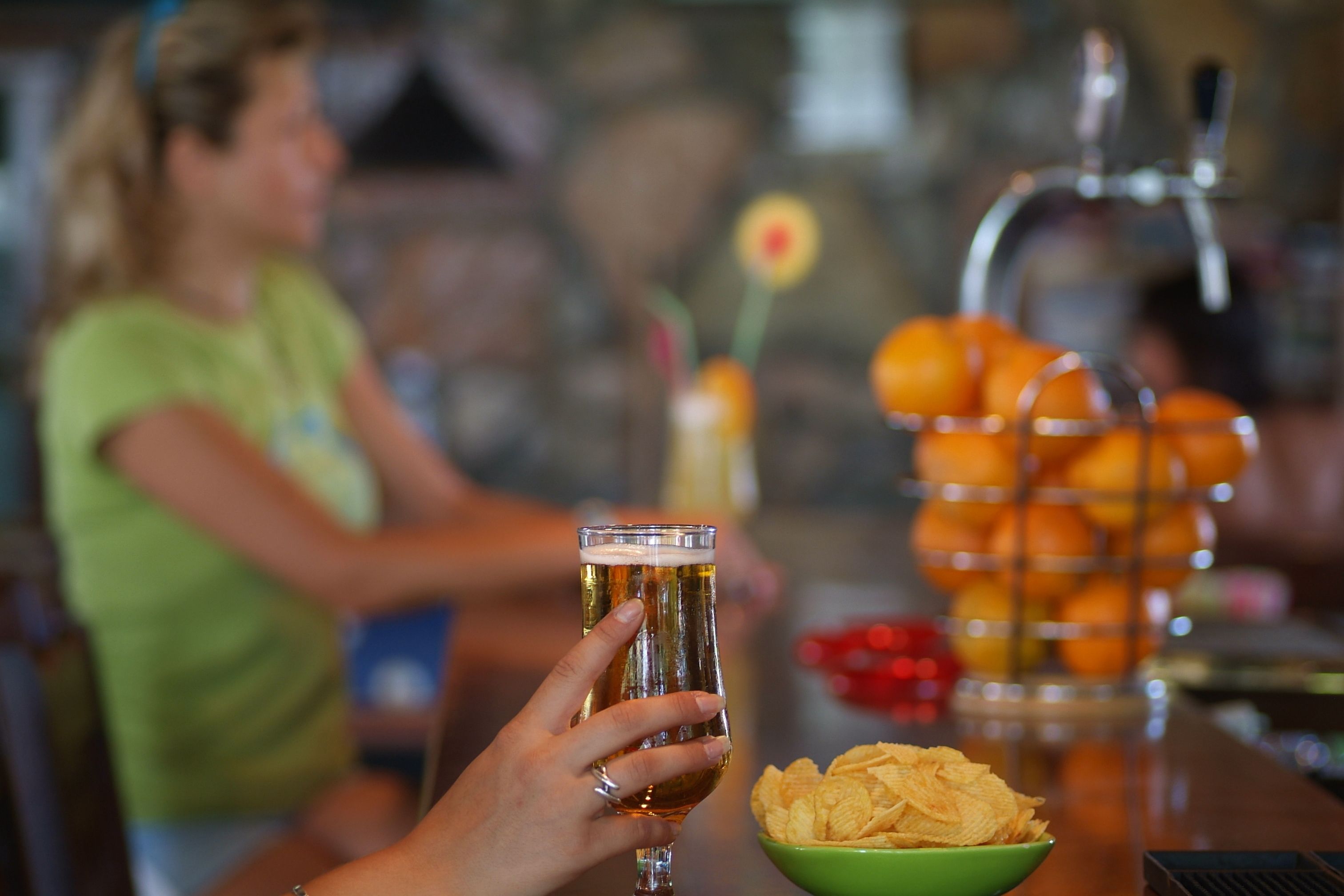En person holder et glas øl i en bar, med en skål chips og en frugtkurv i baggrunden.