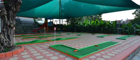 A miniature golf course with green and red playing surfaces, surrounded by a paved area with circular patterns.