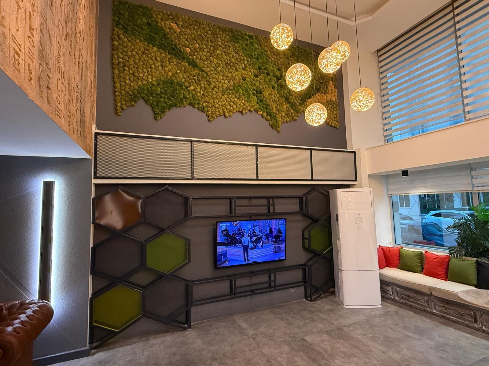 A modern lobby with a large TV, a seating area with colorful cushions, a wall-mounted light fixture, and a green textured wall.
