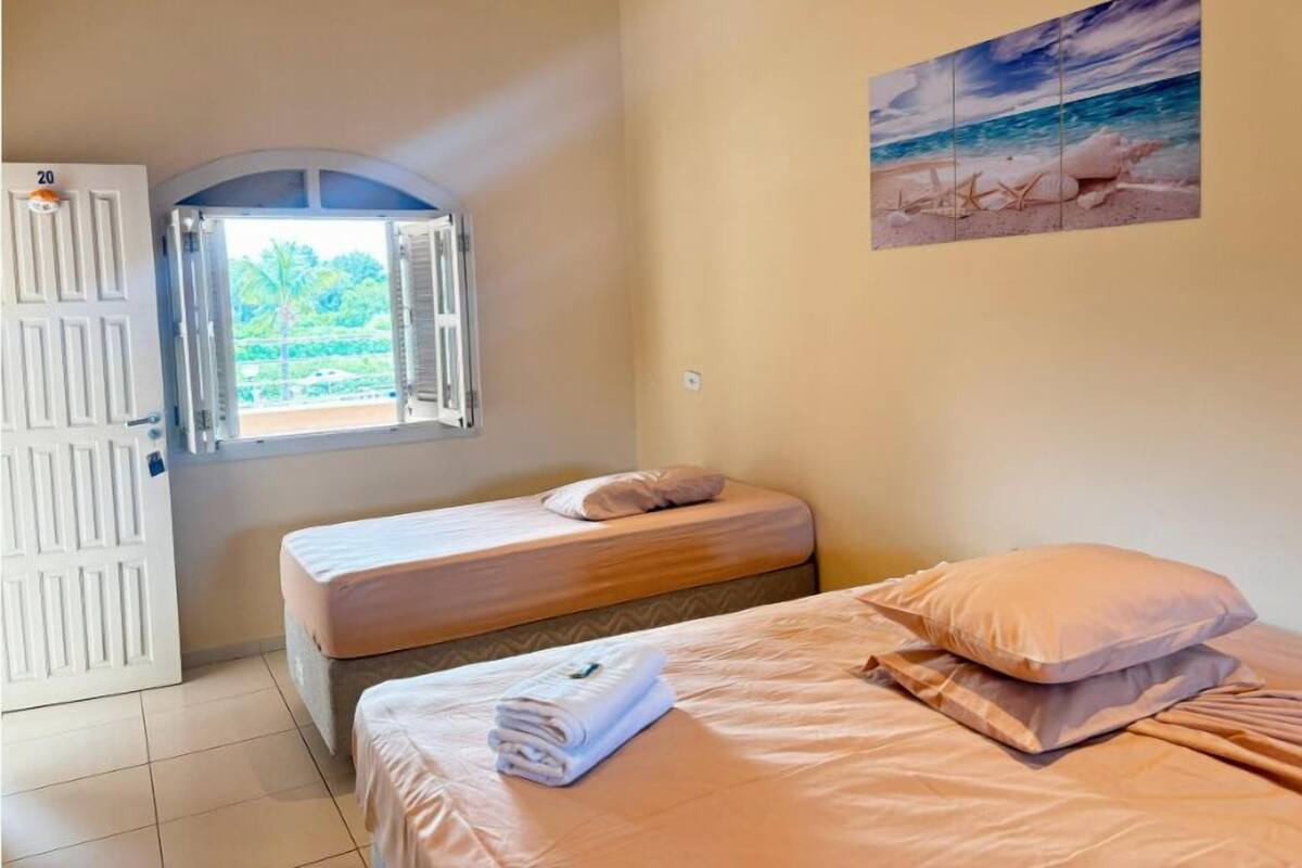 Ein Zimmer mit zwei Betten, einem Fenster mit geöffneten Lamellen, einem Bild von einer Strandansicht und einer Tür mit einer Nummer.