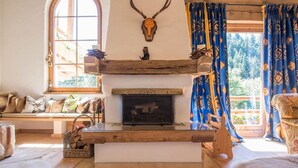 Un salon chaleureux avec une cheminée, des poutres apparentes, un crâne de cerf accroché au mur et une grande fenêtre avec des rideaux.