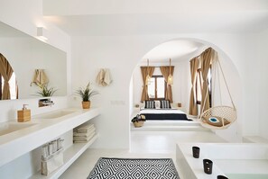 A modern bathroom with a large mirror, white fixtures, a hanging chair, and a bed with a view. - Le Blanc Suite (Santorini)