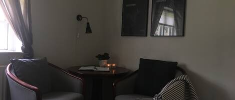 Una habitación pequeña, con poca luz, que tiene dos sillones, una mesita con una planta y dos cuadros en la pared.