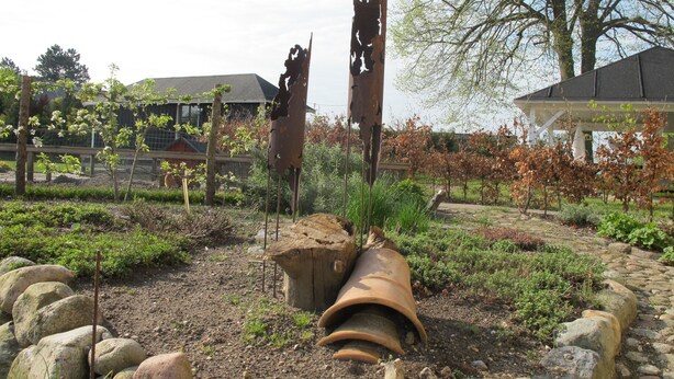 A garden with a rustic, weathered metal sculpture, a large terracotta pot, and a stone pathway. - Birkende Bed And Breakfast (Langeskov)