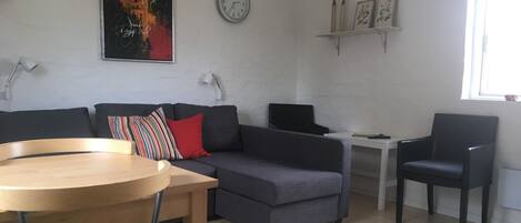 A modern living room with a grey sofa, a wooden table, a black armchair, a white clock, and a framed picture on the wall.