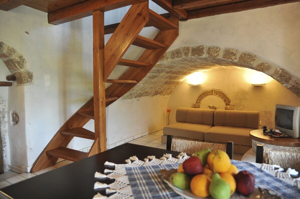 A rustic room with a wooden staircase, a stone wall, a sofa, a television, and a table with fruit.