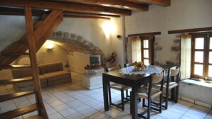 Un salon chaleureux avec un mur en pierre, des poutres apparentes, un canapé, une table avec des chaises et un téléviseur.