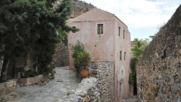 Une ruelle étroite, pavée de pierres, bordée d’un bâtiment rustique avec un mur en pierre et un pot de fleurs aux fleurs rouges.