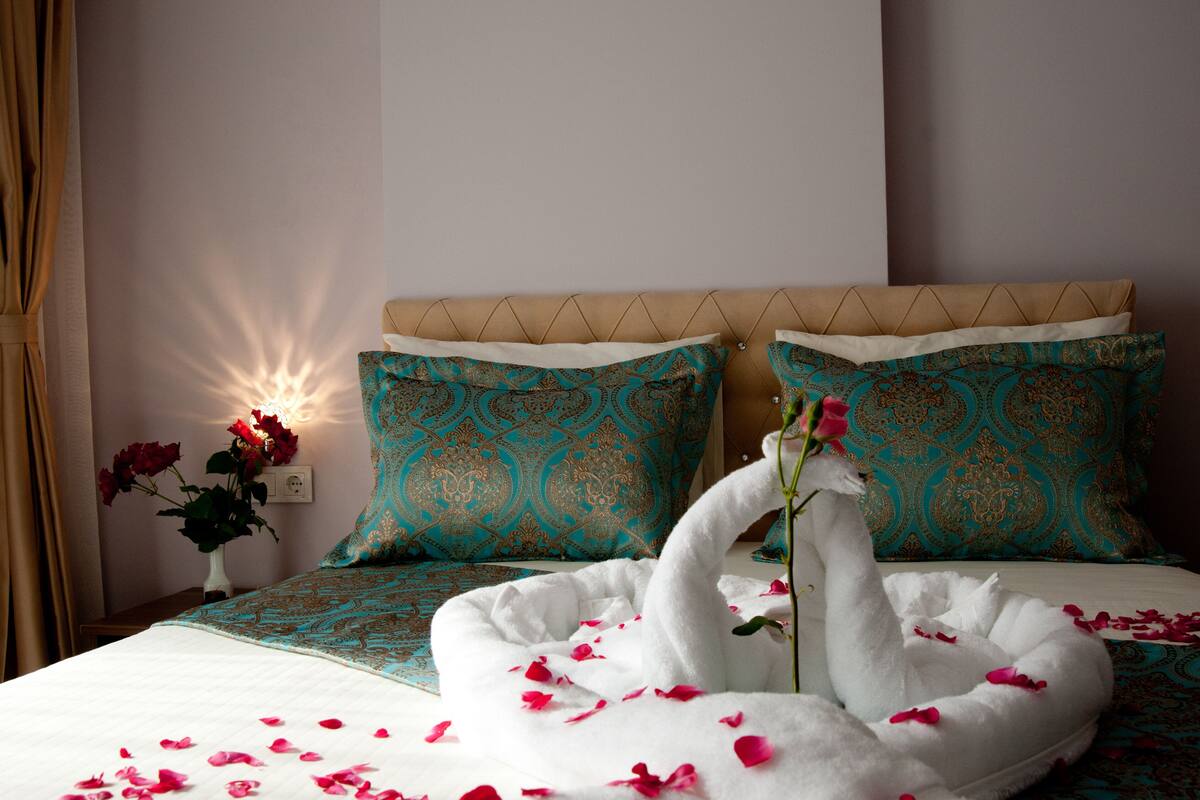 A neatly made bed with a swan-shaped pillow and rose petals, teal patterned pillows, and a vase with red roses on a bedside table.